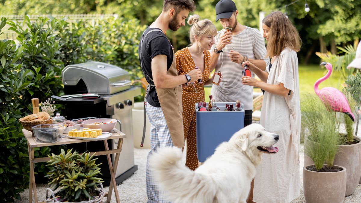 Two men two women and a dog stand by a barbecue in the garden with food ready to cook